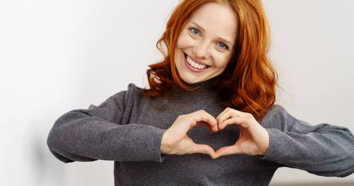 Uśmiechnięta rudowłosa kobieta pokazuje symbol serca ułożony z dłoni